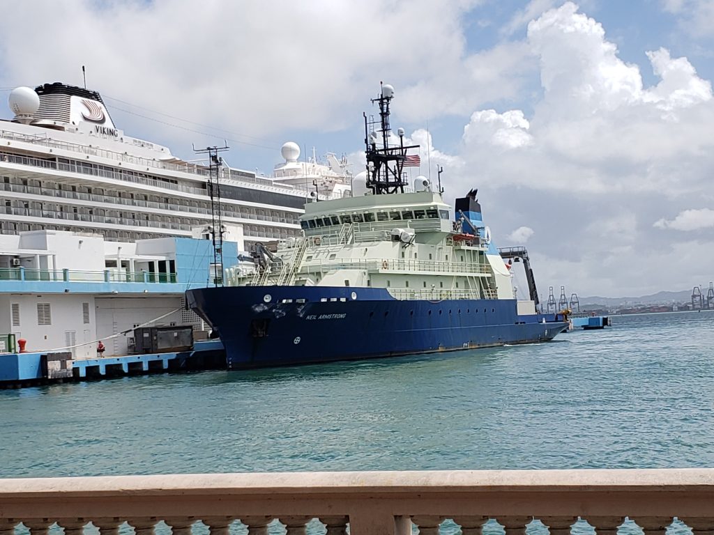 The Research Vessel Neil Armstrong tied up at the dock in San Juan, Puerto Rico. The image is from the waterfront vantage, showing the port side of the vessel. Behind it is a cumulus-cloud sky and a giant cruise ship on the other side of the dock buildings.