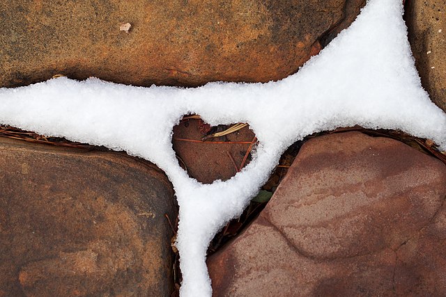 A heart rock in the middle of other rocks with snow surrounding and lining all the cracks.